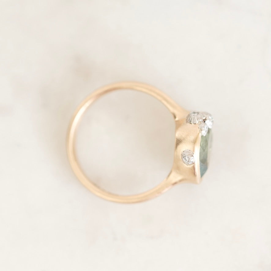 A gold ring with a large green gemstone and small clear accent stones, photographed on a light marble surface.