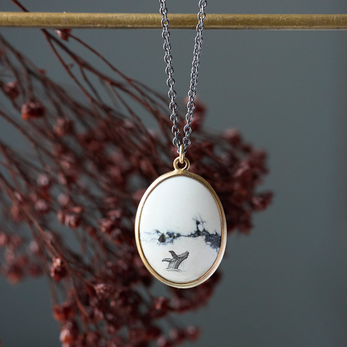 An oval pendant necklace with a white enamel background featuring a black ink-style landscape and bird design, set in a gold-tone frame and hanging from a silver chain. The pendant is displayed against a dark gray background with blurred reddish-brown dried flowers and a gold horizontal bar.