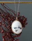 An oval pendant necklace with a white enamel background featuring a black ink-style landscape and bird design, set in a gold-tone frame and hanging from a silver chain. The pendant is displayed against a dark gray background with blurred reddish-brown dried flowers and a gold horizontal bar.