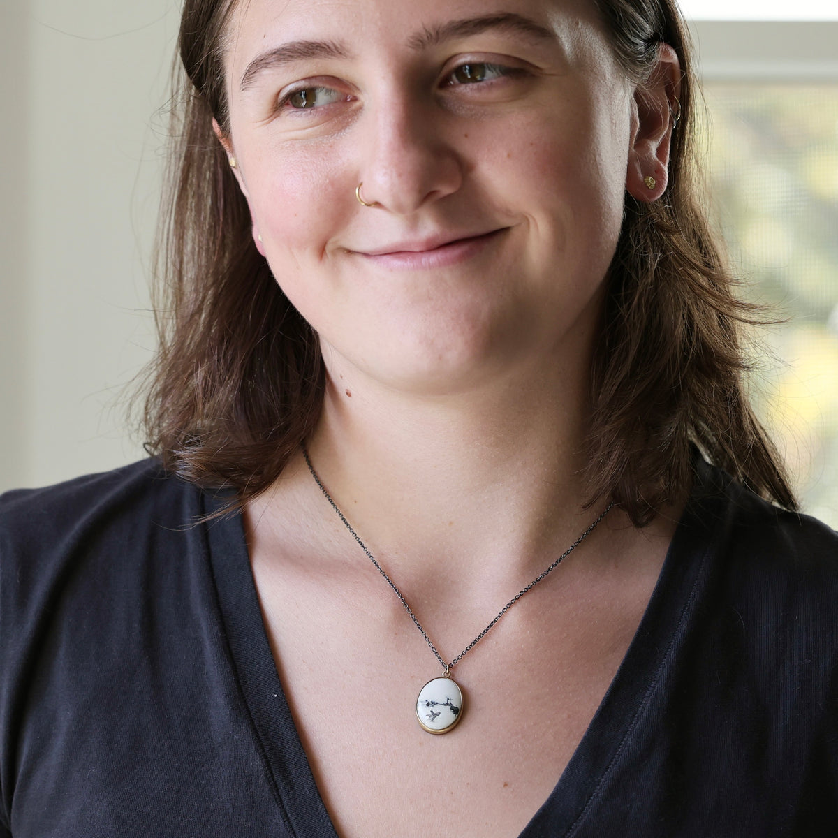A woman wearing a silver chain necklace with a round white pendant engraved with a small black face, shown in a close-up portrait.
