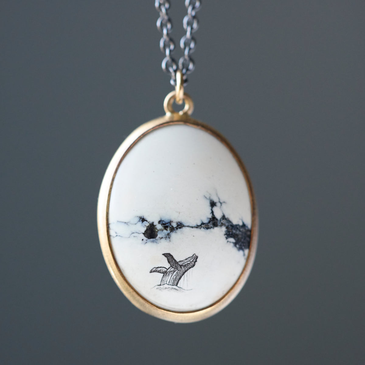 An oval pendant necklace featuring a white stone with black veining and a small whale tail illustration, set in a gold-tone bezel with a silver-tone chain.