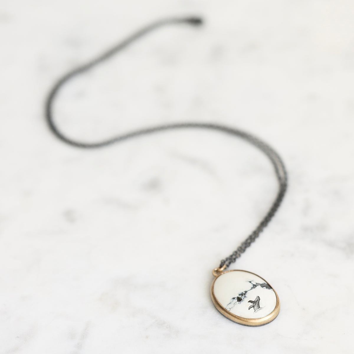 A delicate black cord necklace with an oval pendant featuring a white background, gold-toned border, and black bird illustrations, displayed on a light marble surface.