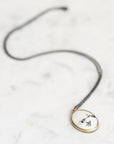 A delicate black cord necklace with an oval pendant featuring a white background, gold-toned border, and black bird illustrations, displayed on a light marble surface.