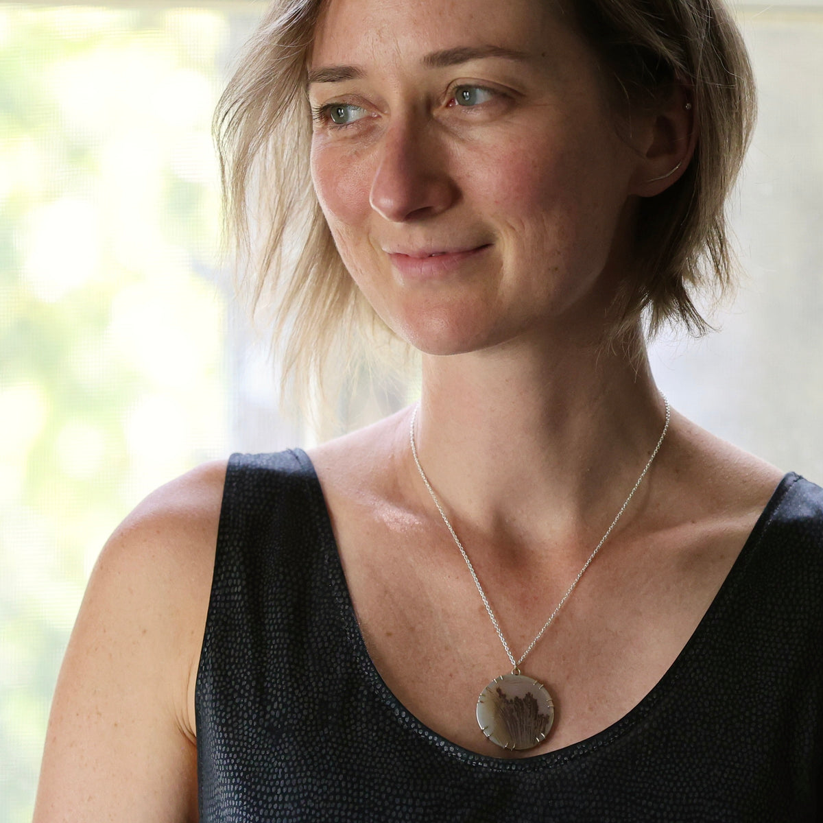 A woman wearing a silver chain necklace with a round, textured stone pendant, photographed in a bright indoor setting.