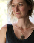 A woman wearing a silver chain necklace with a round, textured stone pendant, photographed in a bright indoor setting.