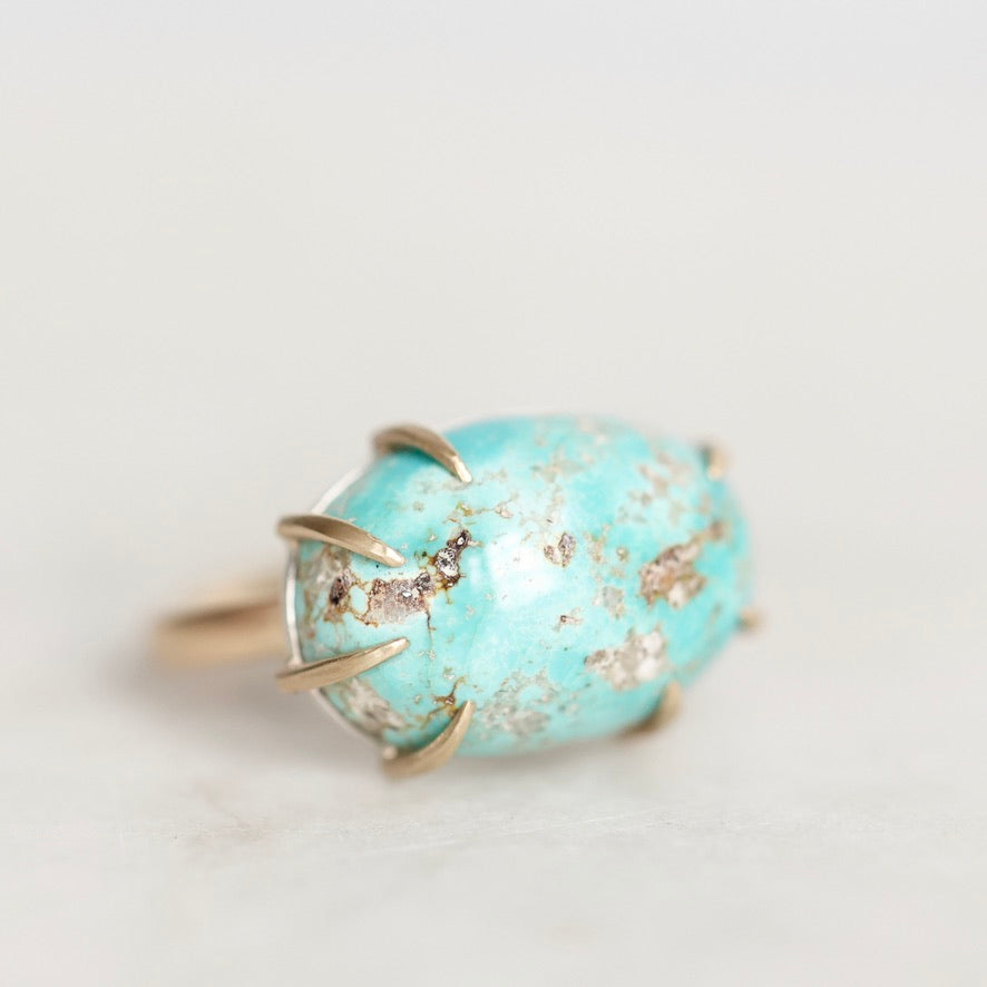A gold ring featuring a large, oval-shaped turquoise stone with natural matrix inclusions, photographed on a clean, light off-white background.