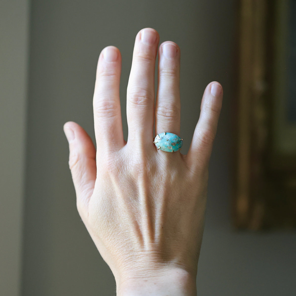 A hand wearing a ring with a turquoise-blue stone set in a silver-toned band, shown against a softly blurred indoor background.