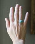 A hand wearing a ring with a turquoise-blue stone set in a silver-toned band, shown against a softly blurred indoor background.