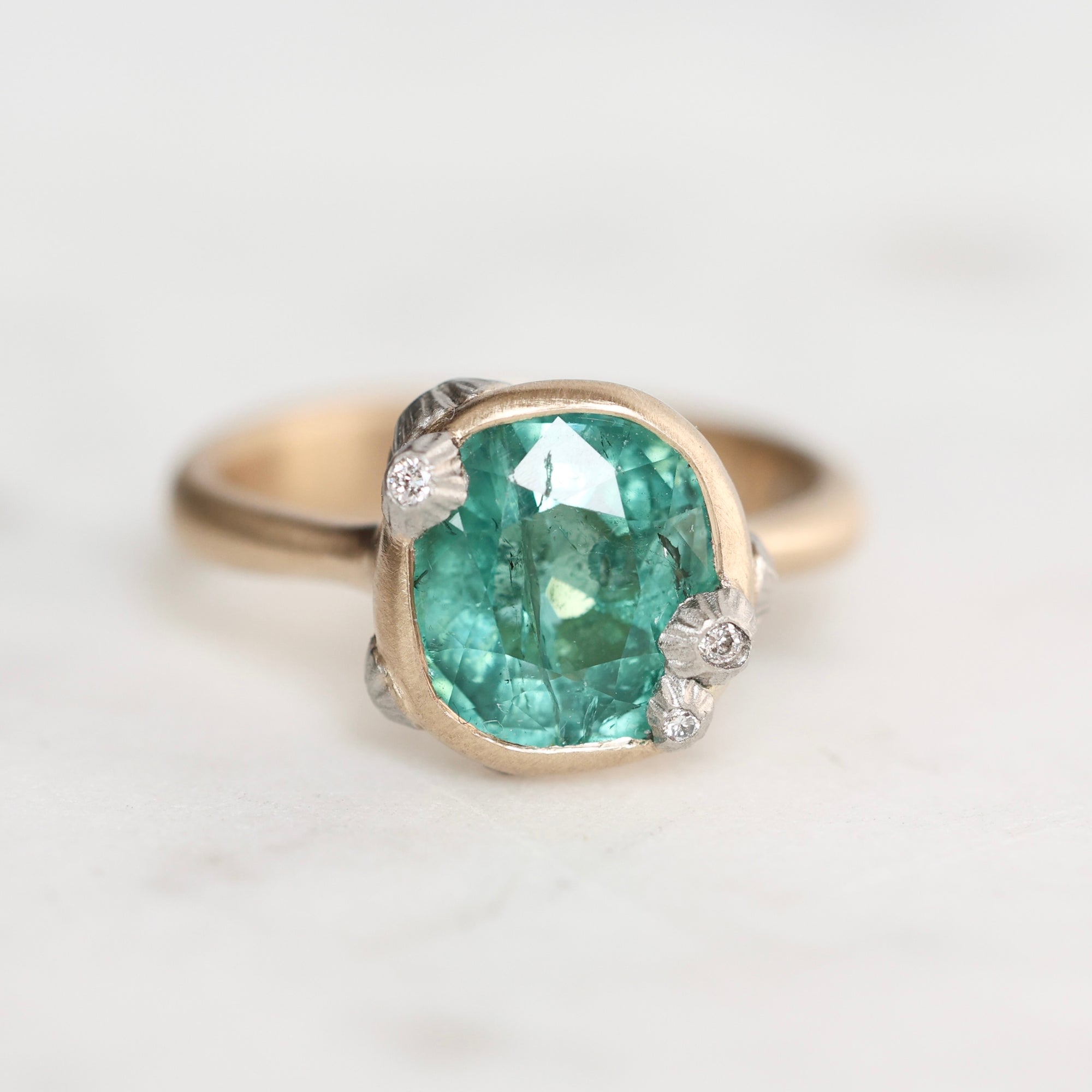 A close-up shot of a ring featuring a cushion-cut green gemstone in a gold bezel setting, accented with small white diamond-like stones on the shoulders, set against a light gray background.