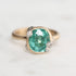 A close-up shot of a ring featuring a cushion-cut green gemstone in a gold bezel setting, accented with small white diamond-like stones on the shoulders, set against a light gray background.