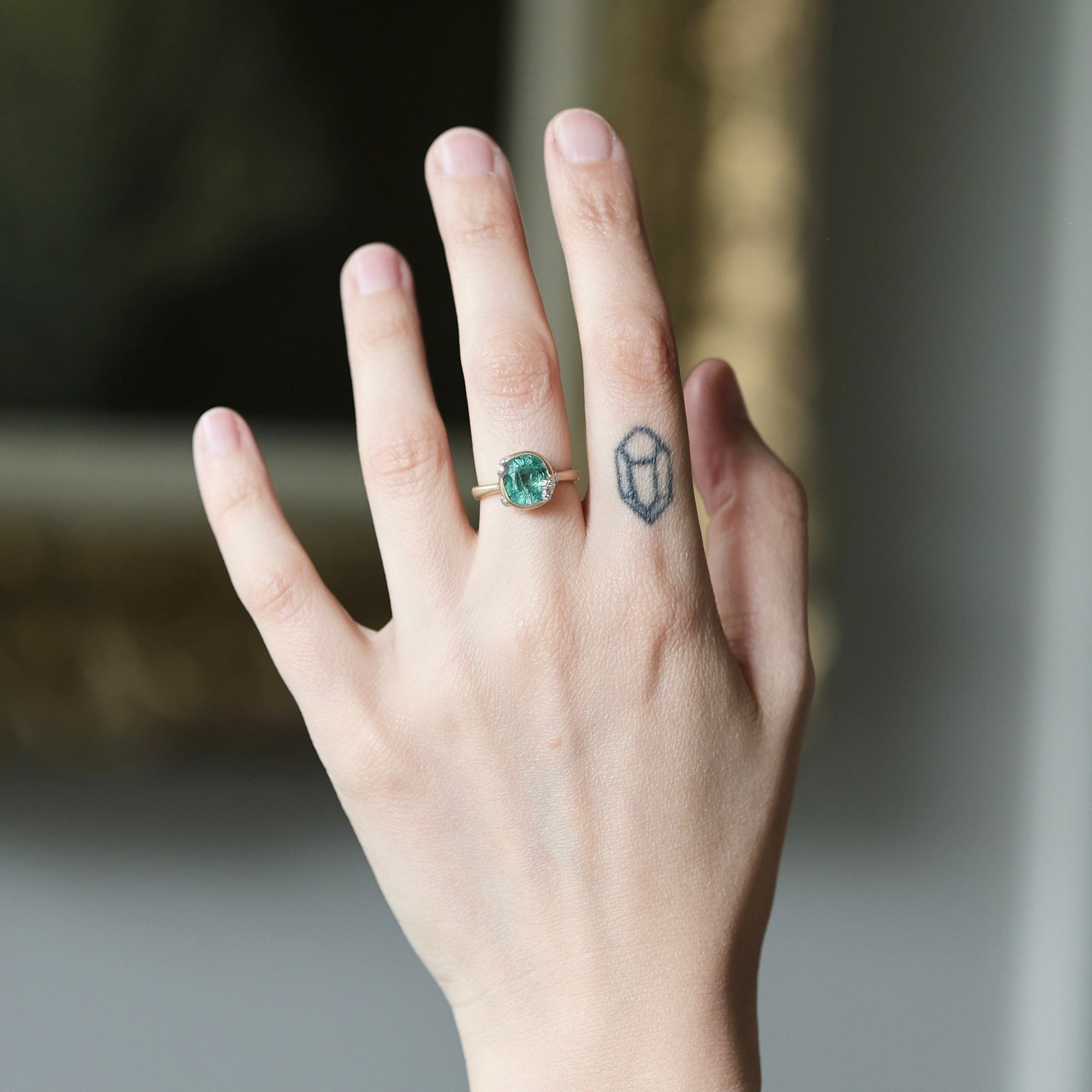 A close-up of a person's hand wearing a gold ring with a round green gemstone, set against a softly blurred indoor background.