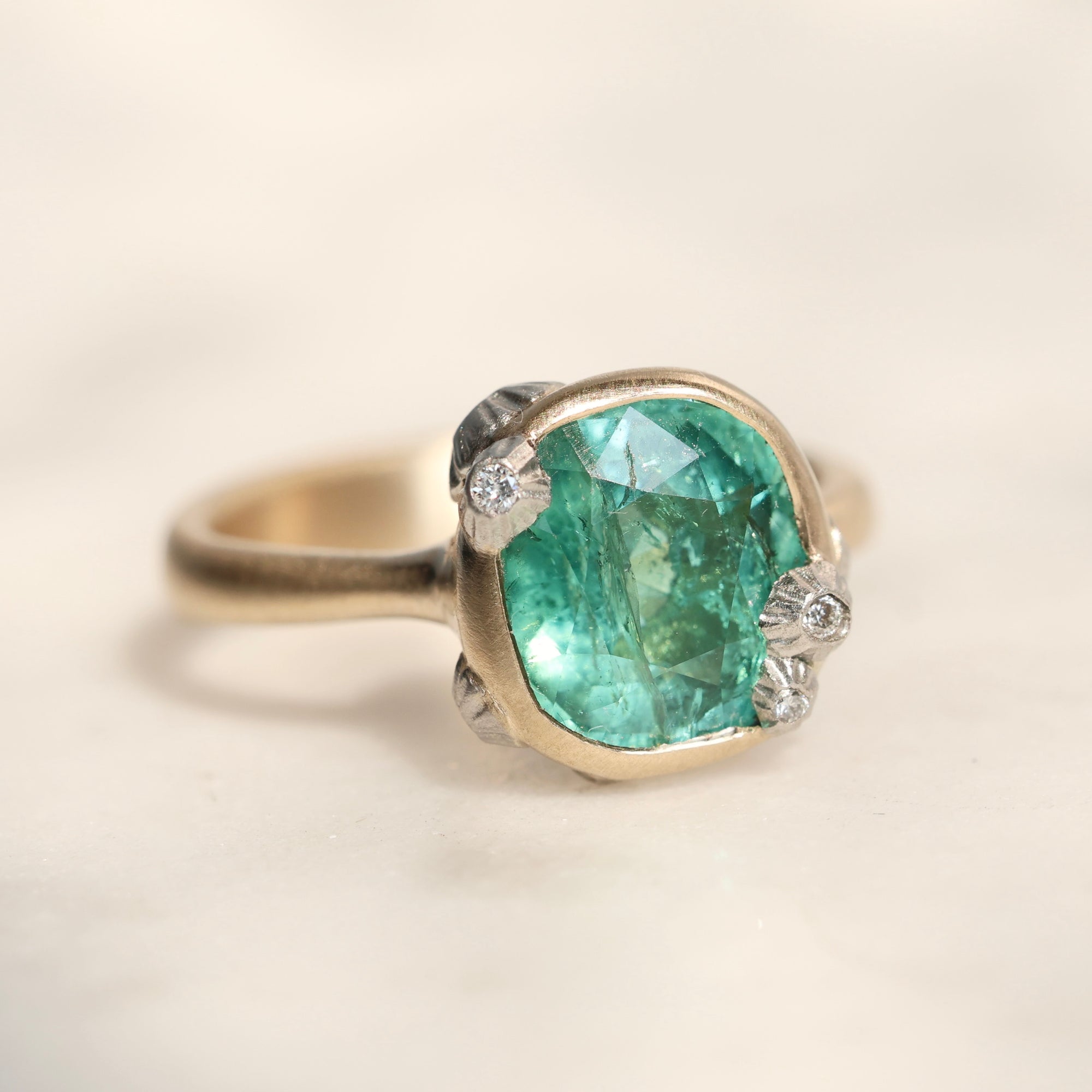 A cushion-cut green gemstone ring set in a gold-toned band with small diamond accents on the sides, photographed on a soft cream background.