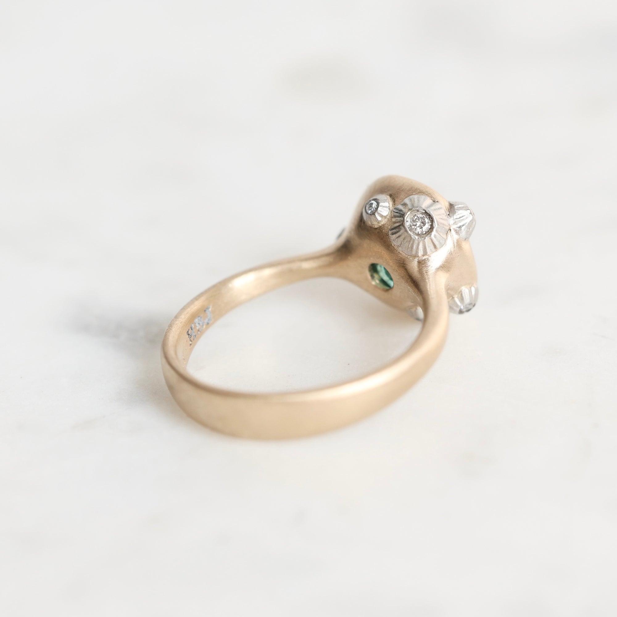 A vintage-style ring with a gold band and a silver-toned decorative top featuring a central round diamond, smaller side stones, and a tiny green accent, photographed on a light marble surface.
