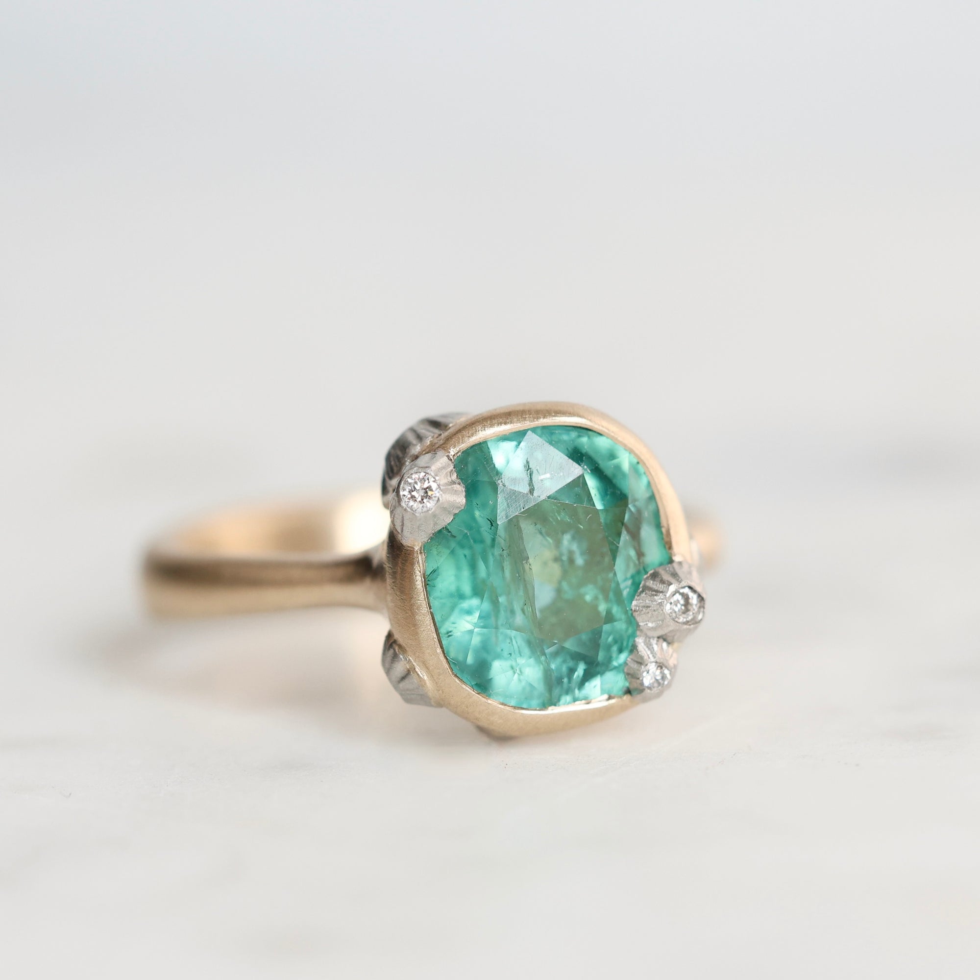 A cushion-cut green gemstone ring set in a gold-toned band with small clear accent stones, photographed on a light neutral background.