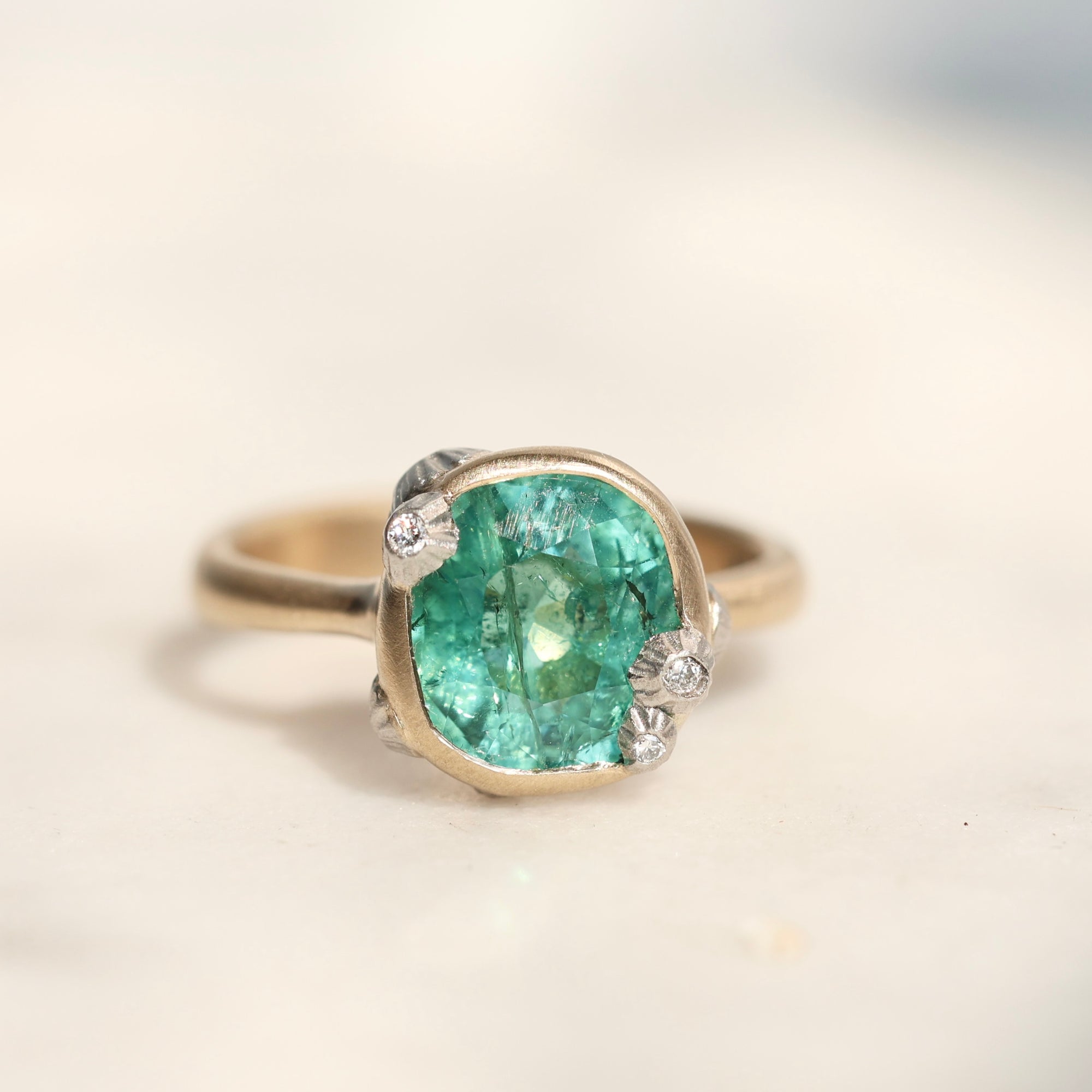 A vintage-style ring featuring a large cushion-cut green gemstone in a gold-toned setting with small clear accent stones, photographed on a light beige surface.