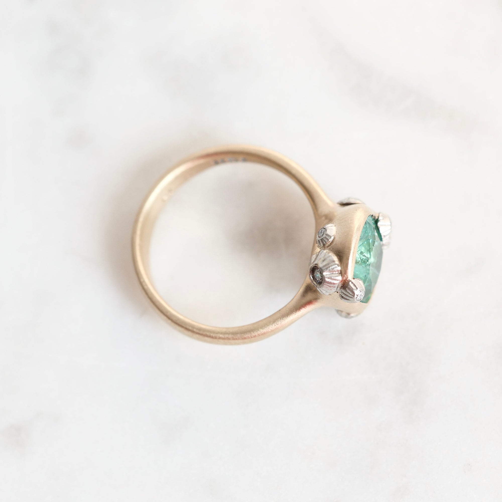 A close-up shot of a vintage-style ring with a rose gold band, featuring a green  gemstone and small silver floral accents, resting on a white marble surface.
