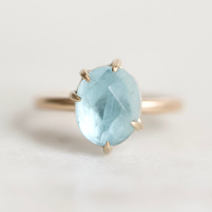 A gold ring featuring an oval-cut light blue gemstone in a four-prong setting, photographed on a clean, light off-white background.