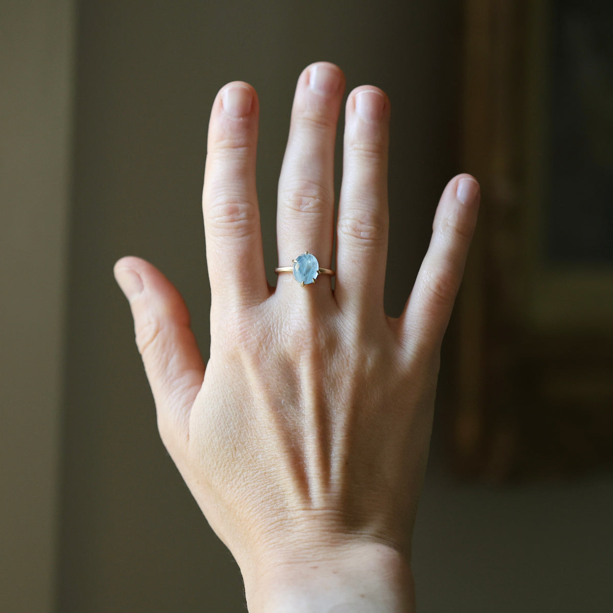 A close-up of a person's hand wearing a delicate gold ring with a light blue oval gemstone, set against a softly blurred indoor background.