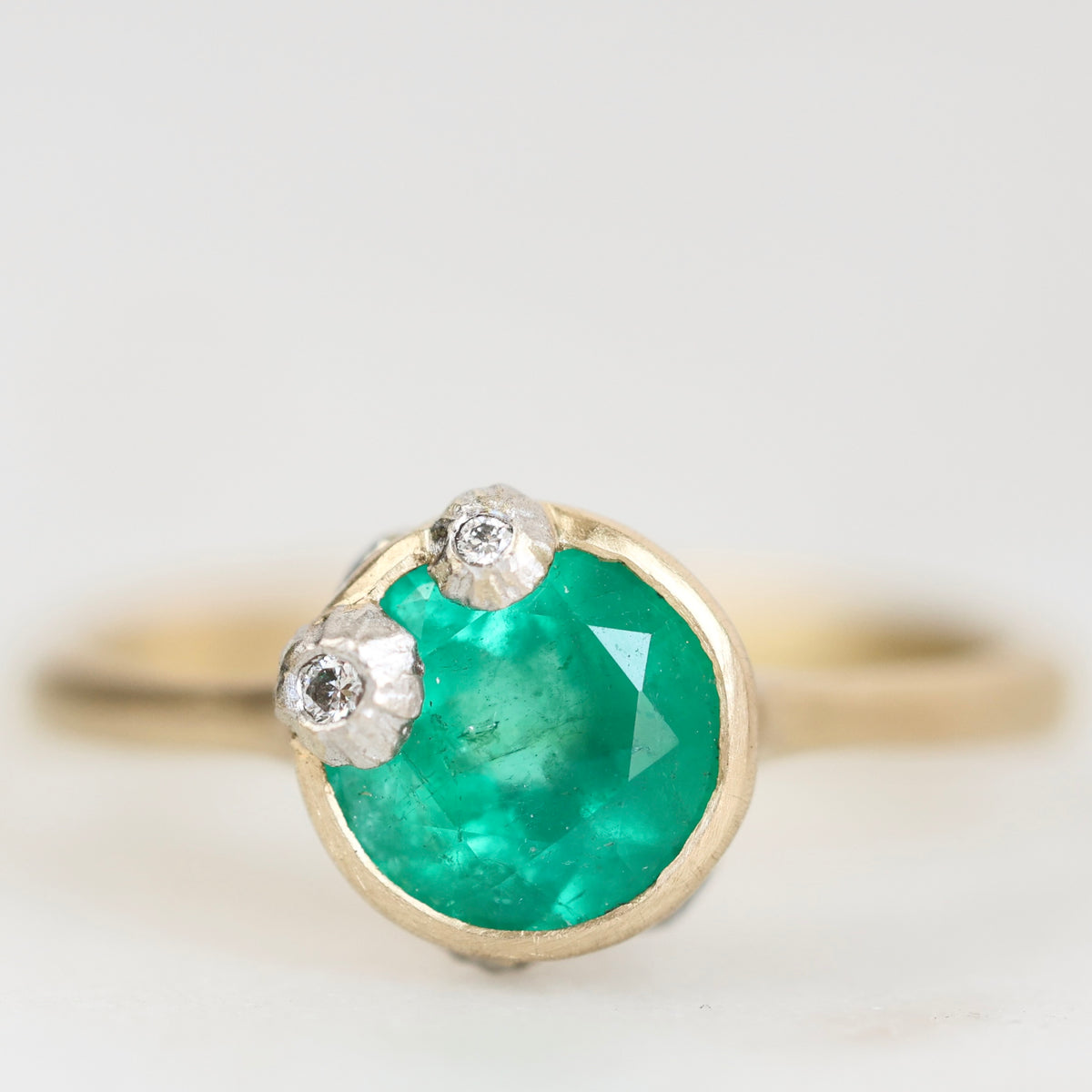 A gold ring featuring a round green emerald-cut center stone and two small silver floral accents, photographed against a soft white background.