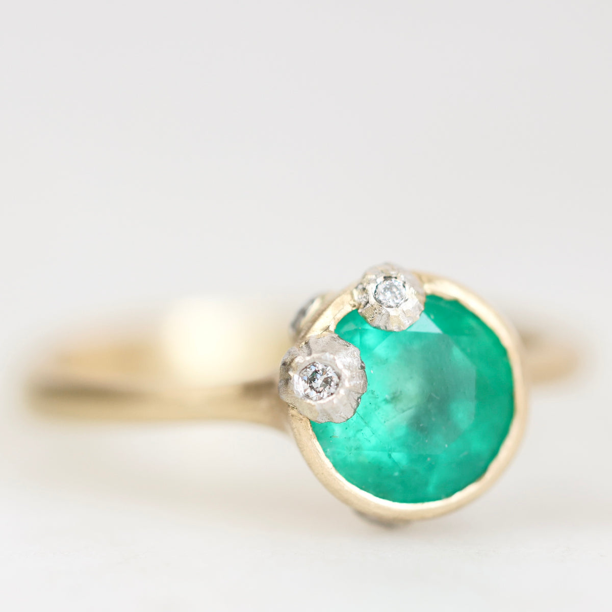 A gold ring featuring a round green emerald-cut gemstone and two small clear diamond accents on the top, shown against a soft white background.