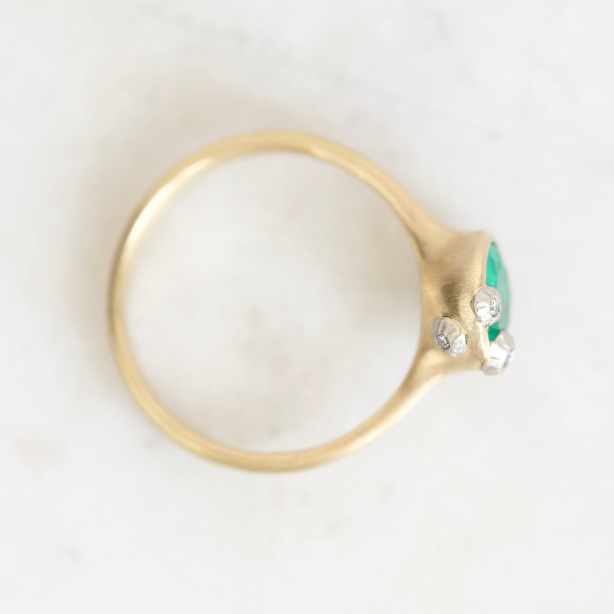 A gold ring with a green marquise-cut center stone and three small clear accent stones, photographed from above on a light marble surface.