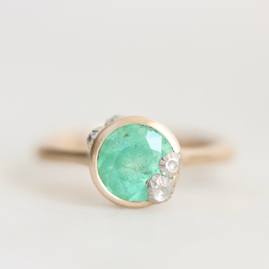 A gold ring featuring a round faceted green gemstone with small clear accent stones on the right side, set against a soft light gray background.
