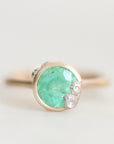 A gold ring featuring a round faceted green gemstone with small clear accent stones on the right side, set against a soft light gray background.