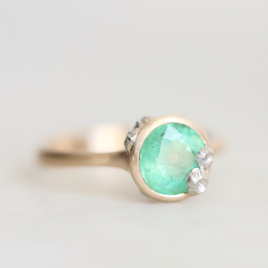A close-up shot of a ring featuring a round, faceted light green gemstone in a gold-toned bezel setting, accented with small platinum barnacles on the sides, set against a soft, light gray background.