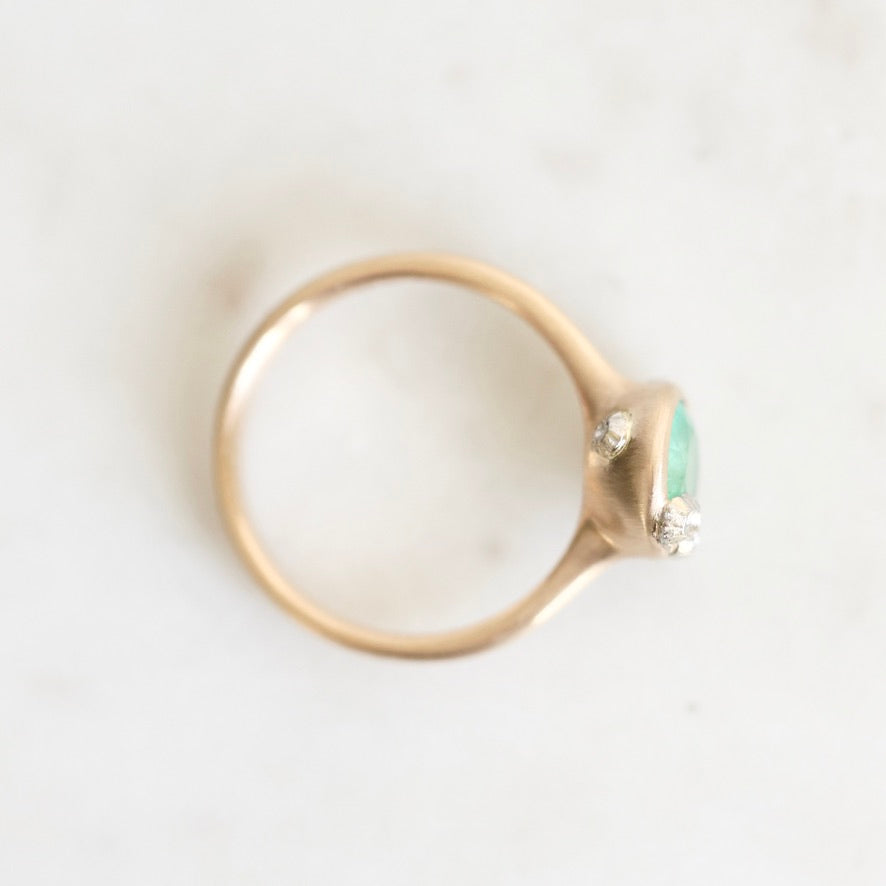 A gold ring with a small green oval gemstone and two accent barnacles, photographed on a light marble-like surface.