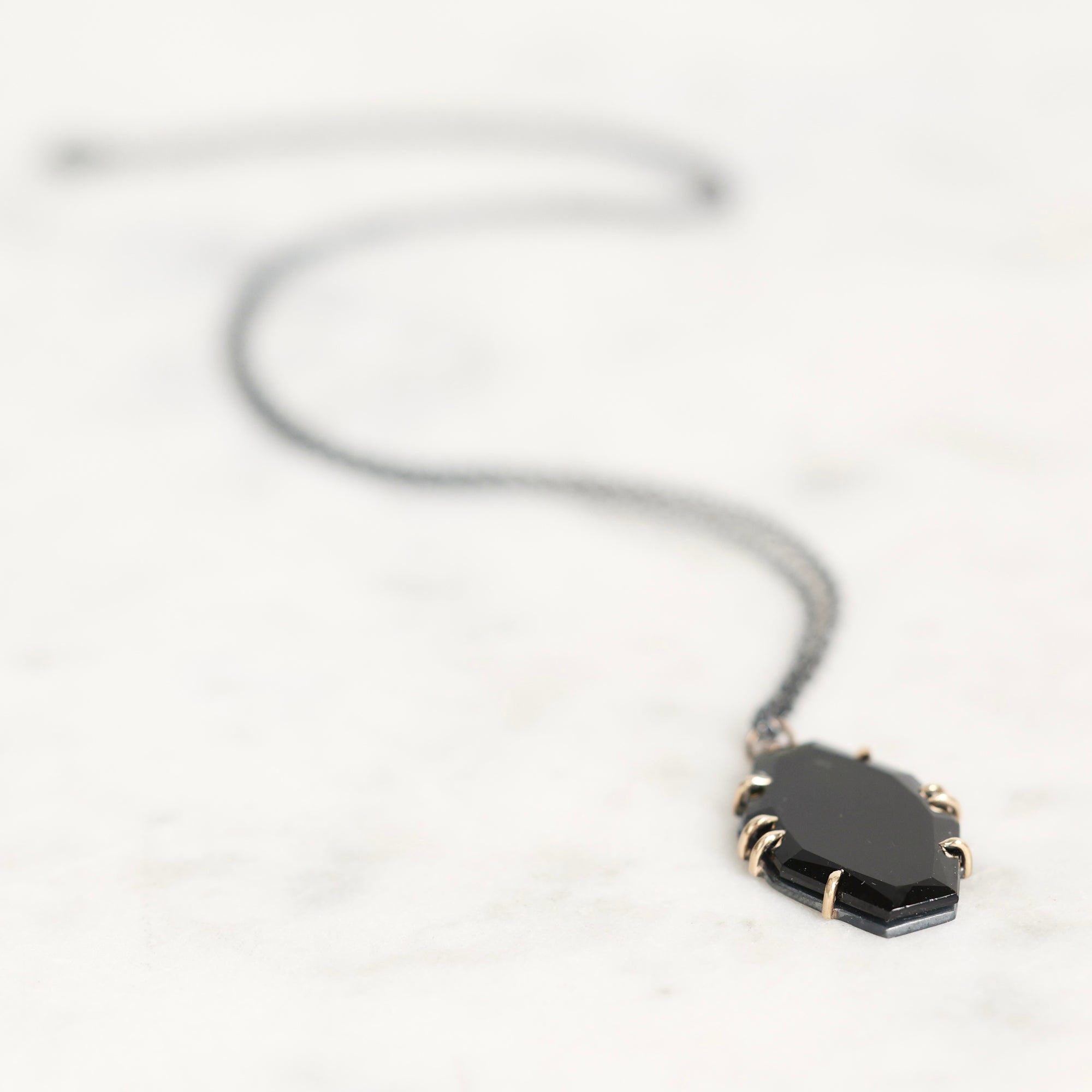A black pendant necklace with a faceted geometric design, held in gold-toned prong settings on a delicate chain, resting on a light marble surface.