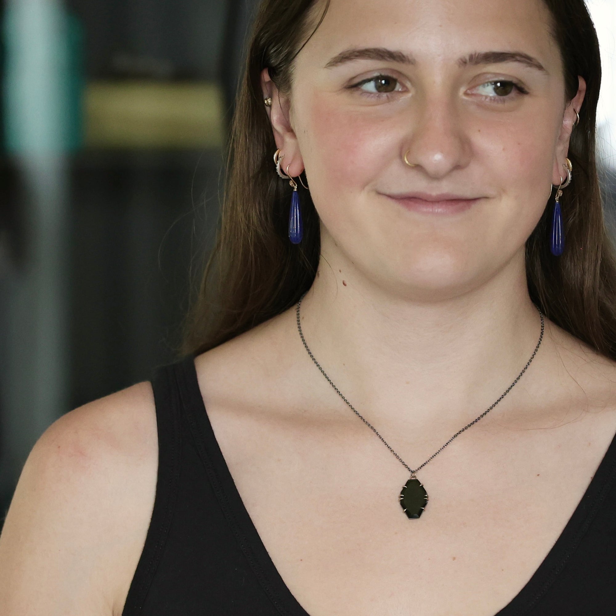 A woman wearing a dark metal pendant necklace and matching drop earrings with purple tassel charms, shown in a close-up portrait.