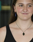 A woman wearing a dark metal pendant necklace and matching drop earrings with purple tassel charms, shown in a close-up portrait.