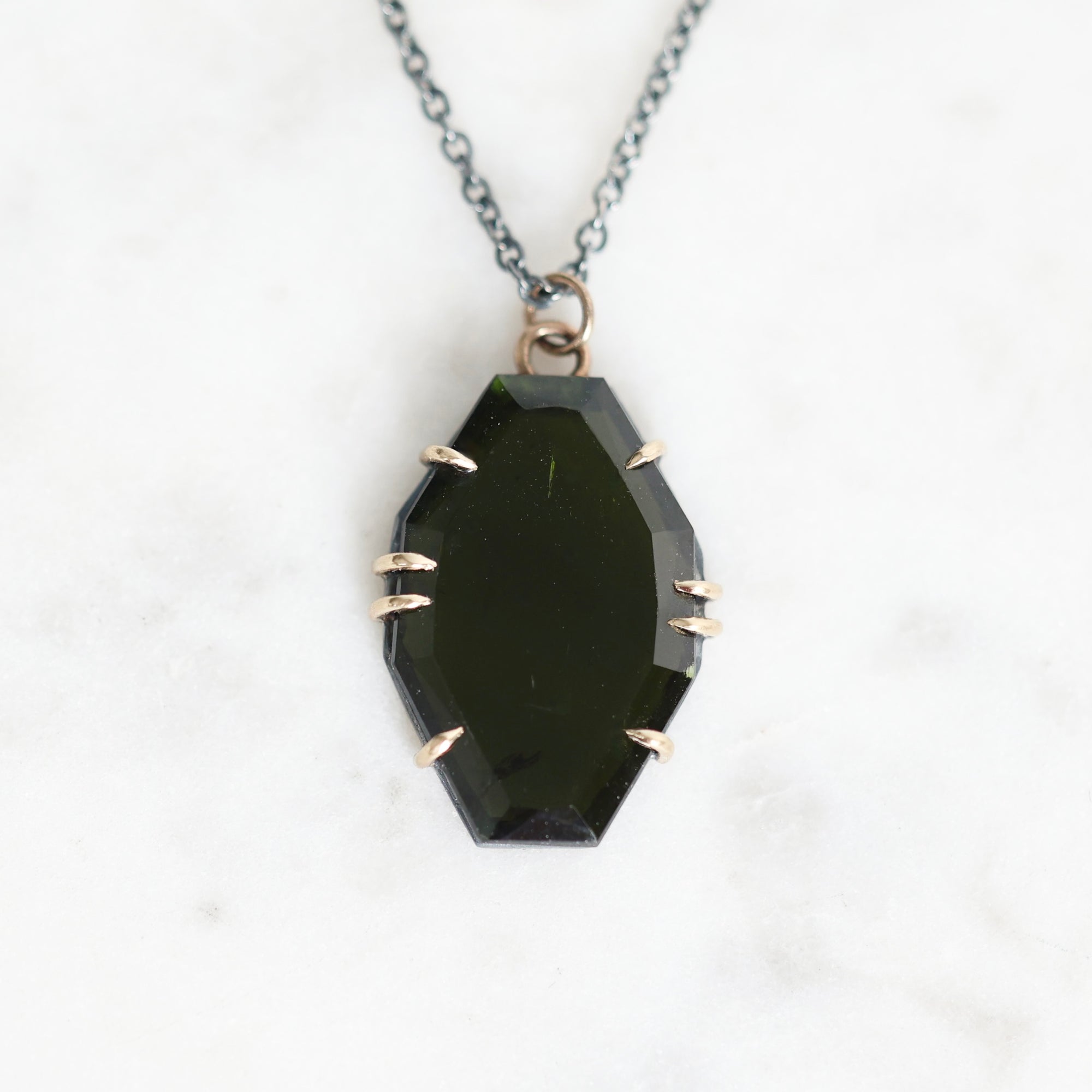 A faceted dark green gemstone pendant necklace with a silver-toned chain and bezel setting, displayed on a white marble background.