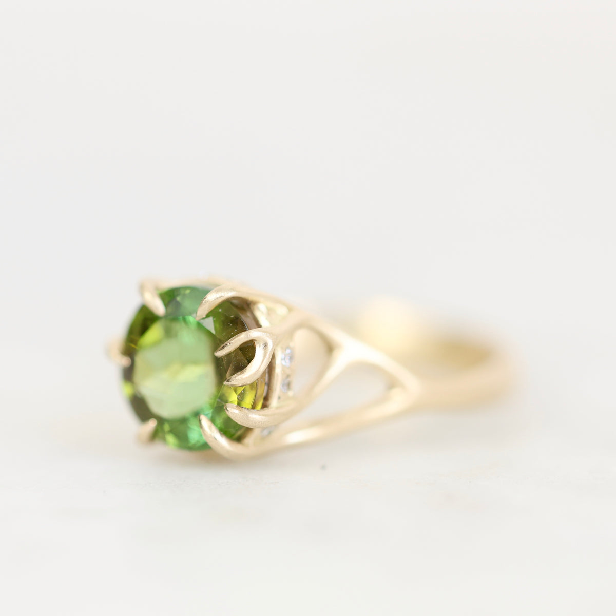 A yellow gold ring featuring a round green peridot gemstone in a prong setting, with a small clear accent stone on the band, shown against a plain white background.