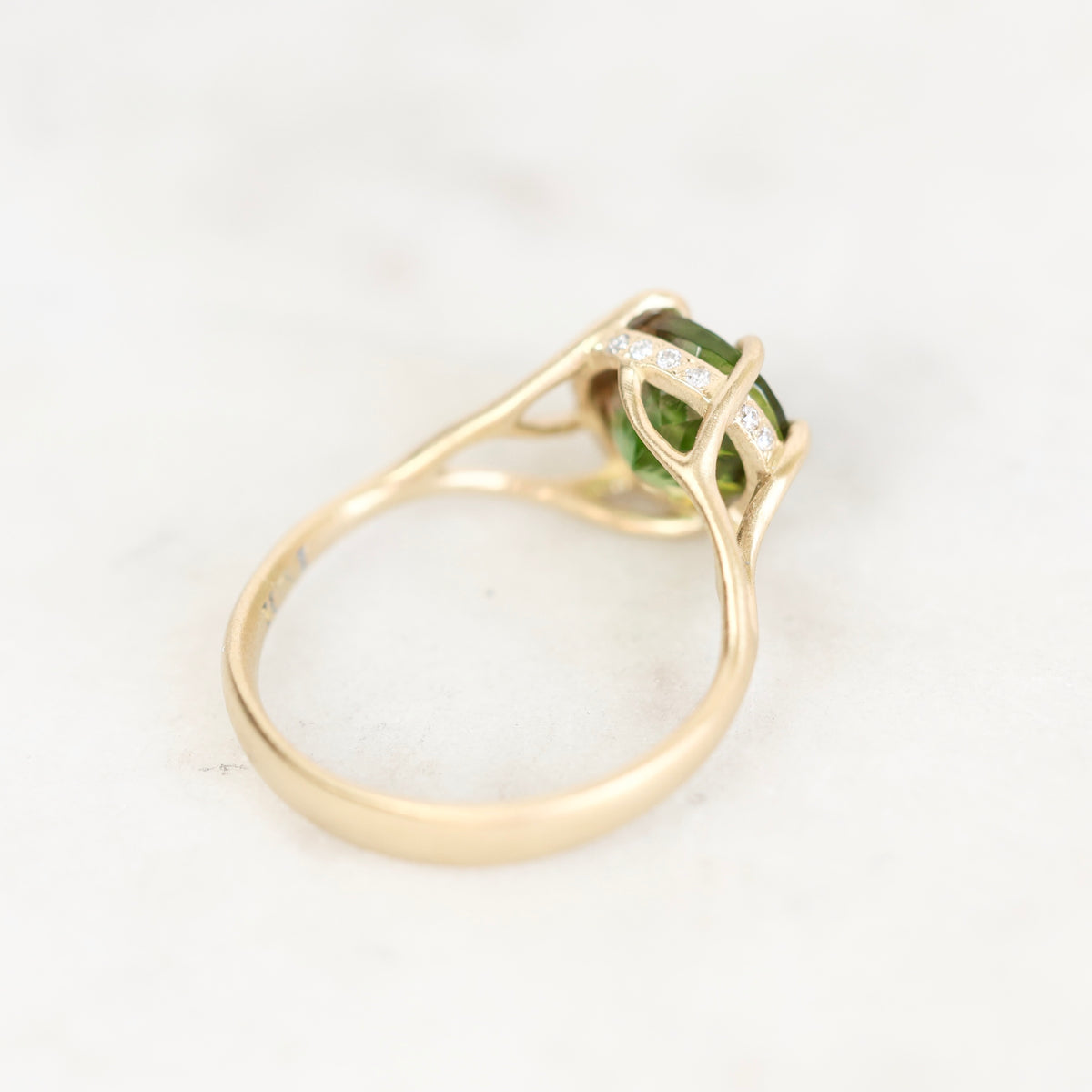 A yellow gold ring featuring a green rectangular gemstone in a prong setting with small clear accent stones, shown on a light marble background.