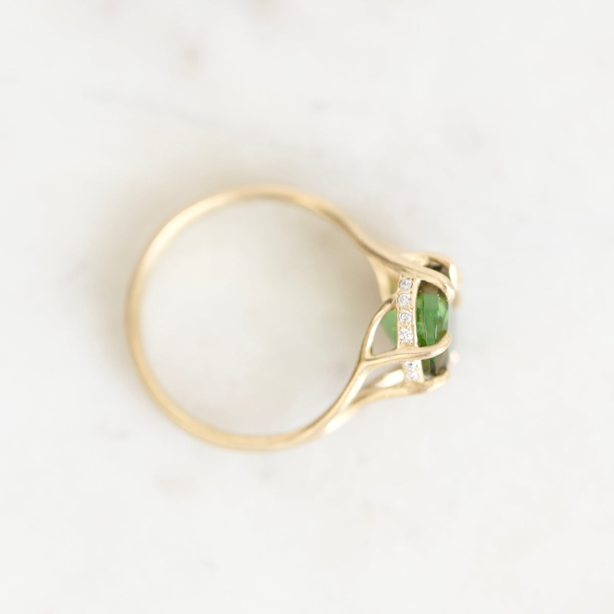 A yellow gold ring with a green oval gemstone and small clear accent stones, photographed on a light marble-like surface.