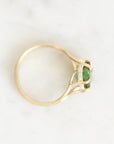 A yellow gold ring with a green oval gemstone and small clear accent stones, photographed on a light marble-like surface.