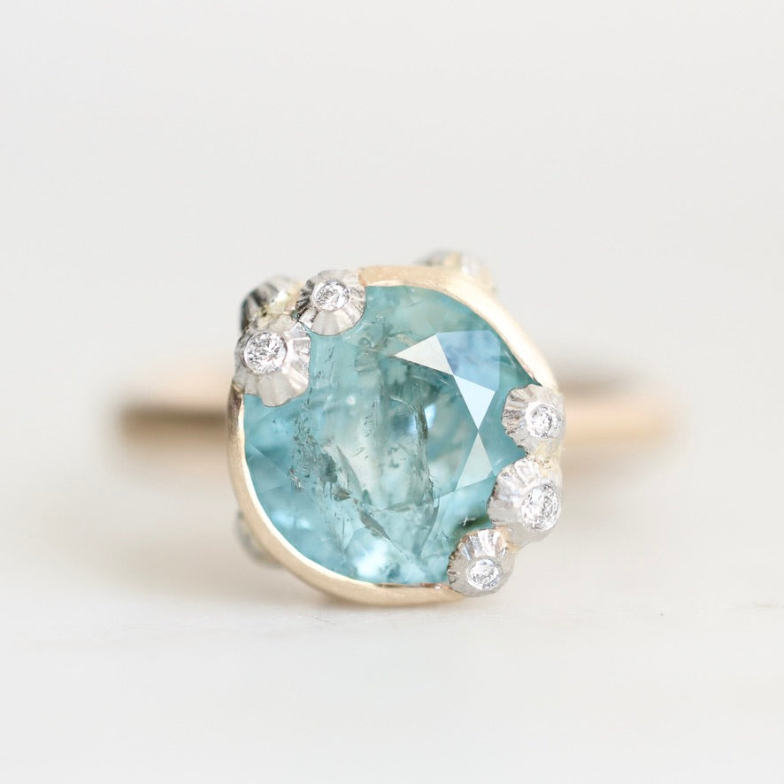 A close-up shot of a ring featuring a large, faceted light blue gemstone in a gold-toned setting, accented by several small round clear stones, set against a plain light background.