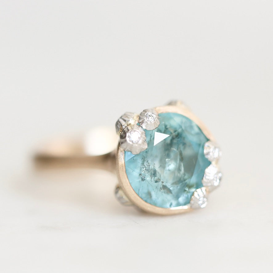 A close-up shot of a ring featuring a large oval light blue gemstone in a gold organic bezel accented with small accent barnacles, on a light gray background.
