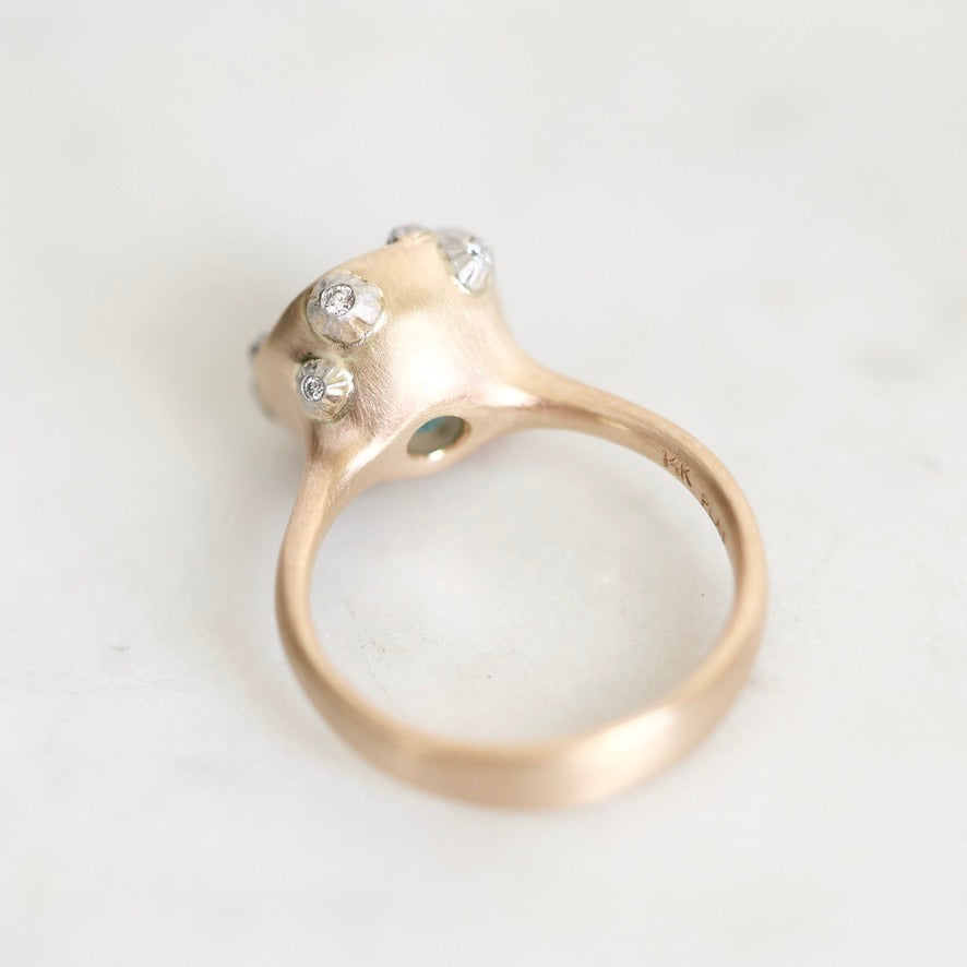 A close-up shot of a gold ring with a textured, organic-shaped setting featuring three small clear gemstones, resting on a light marble surface.
