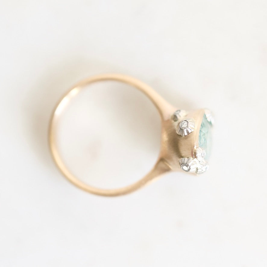 A close-up shot of a ring featuring a gold band and a setting with a light blue-green stone and small clear accent stones, photographed on a plain white background.