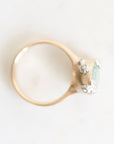 A close-up shot of a ring featuring a gold band and a setting with a light blue-green stone and small clear accent stones, photographed on a plain white background.