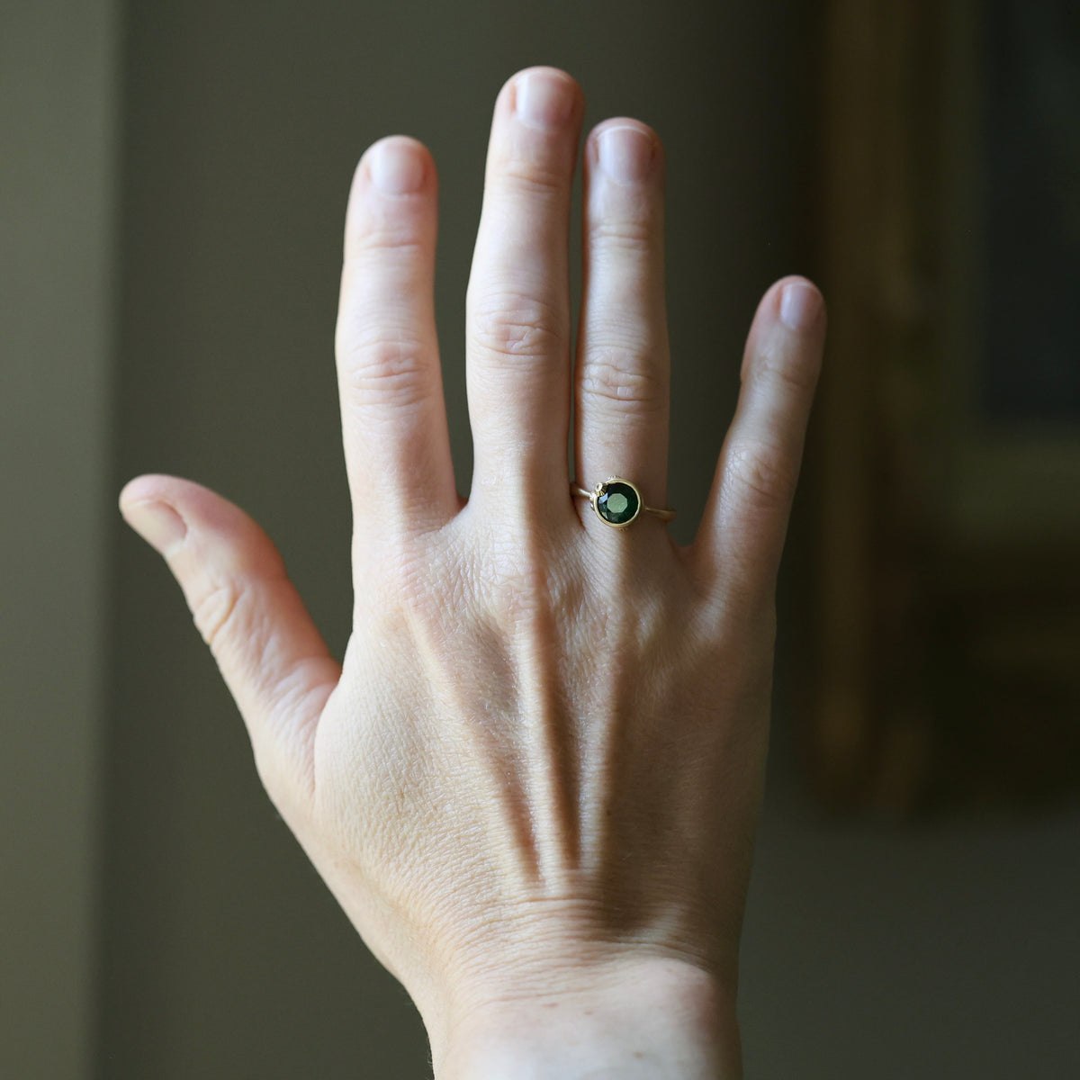 A close-up of a person's hand wearing a gold ring with a round green gemstone, set against a softly blurred indoor background.