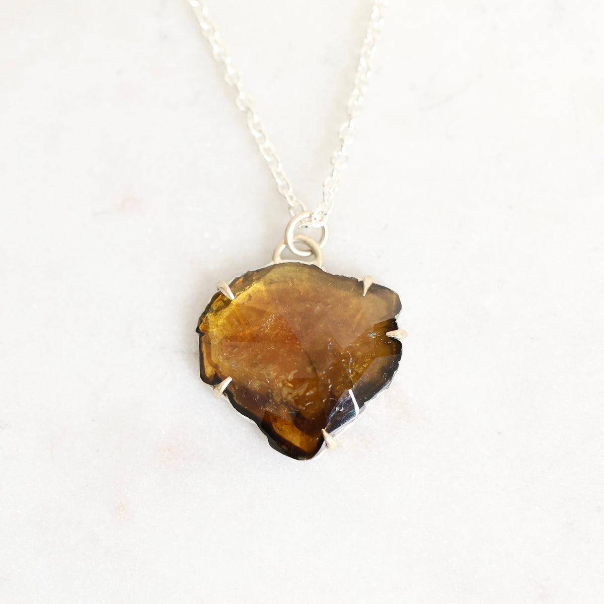 A silver necklace featuring a raw, irregularly shaped amber-brown gemstone pendant, displayed on a clean white background.