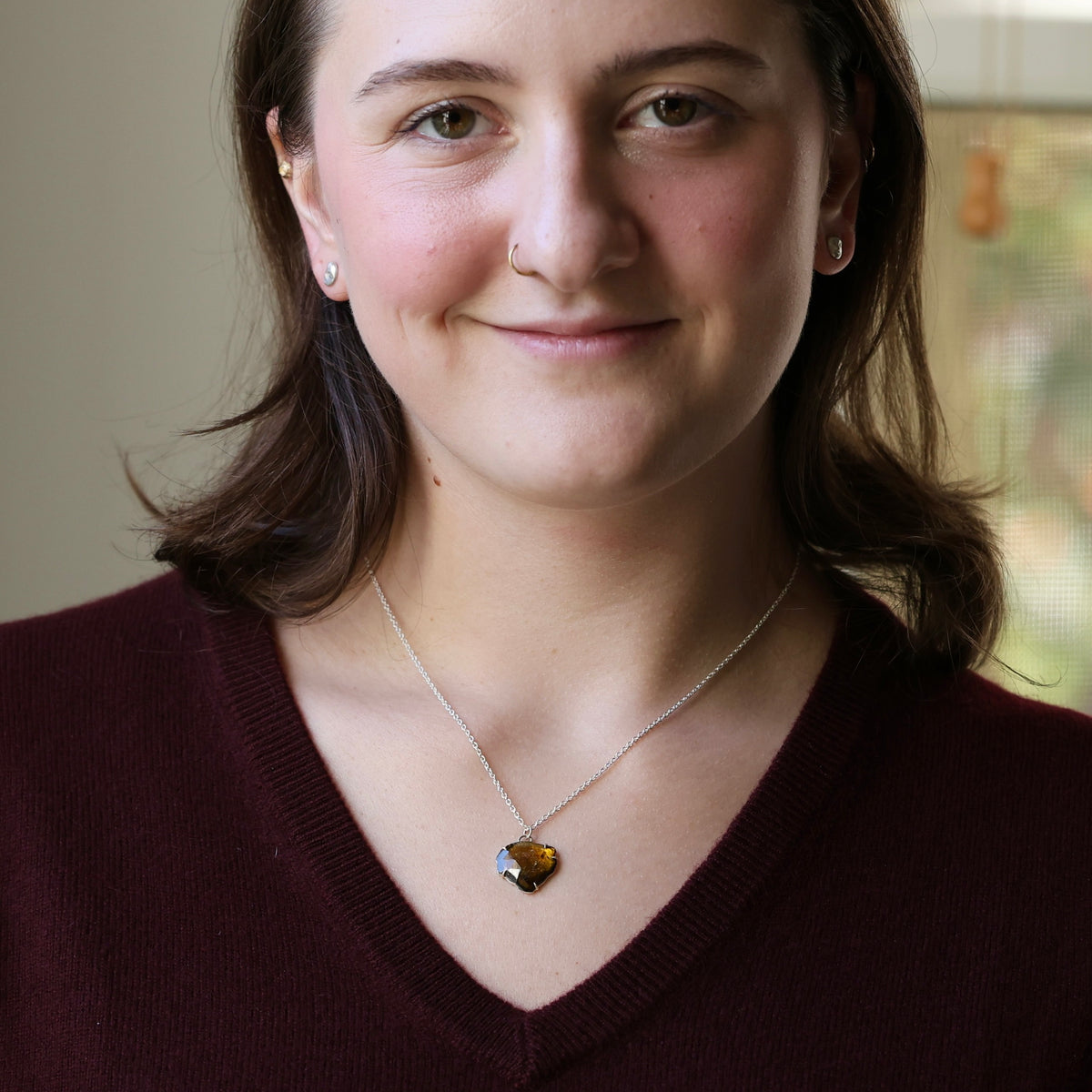 A woman with brown hair wearing a silver chain necklace with a heart-shaped amber gemstone pendant, paired with a burgundy V-neck sweater.