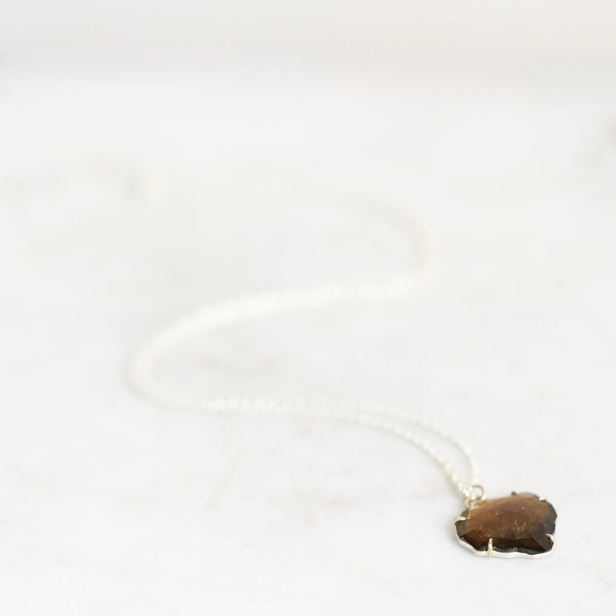 A delicate silver chain necklace with a small, irregular-shaped brown gemstone pendant, resting on a light marble surface.