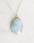 An oval pendant necklace featuring a light blue marbled stone in a gold bezel setting, suspended from a delicate silver-toned chain against a light beige marble background.