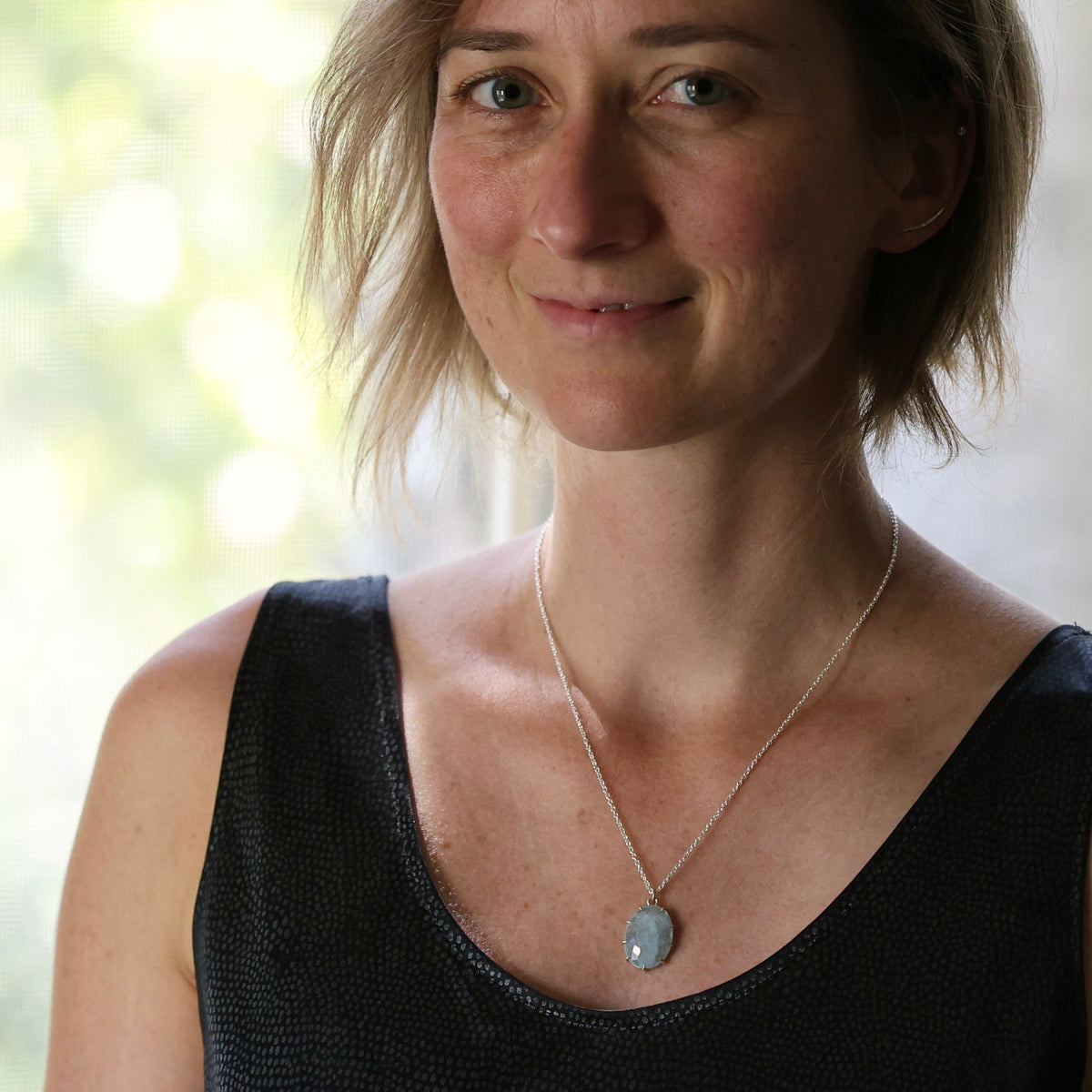 A woman with short blonde hair wearing a silver chain necklace with a light blue-gray oval pendant, paired with a black textured tank top.