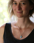 A woman with short blonde hair wearing a silver chain necklace with a light blue-gray oval pendant, paired with a black textured tank top.
