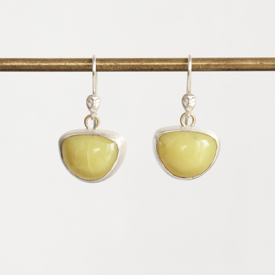 A pair of silver drop earrings with smooth, polished light yellow-green oval stones, hanging from a horizontal brass rod against a light gray background.